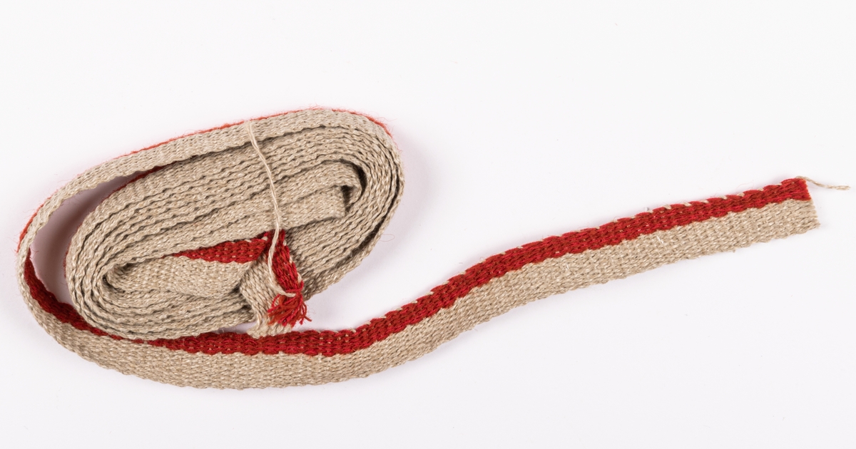 Vävt band, kantband till klänning, ur materialsats till hembygdsdräkt, den så kallade Hoforsdräkten. Oblekt lin och rött garn.