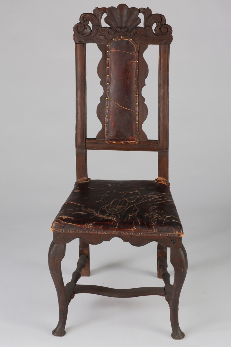 Høyrygget barokkstol med skinnsete og skinntrukket ryggfjøl. Stolen har et toppstykke med kronet akantus. Stolbenene foran er svungne og de bakerste er rette, benene er satt i et H-kryss.