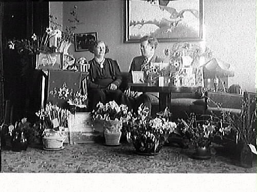 Födelsedagsfirande hos gelbgjutare Karl Holmkvist med fru. De sitter i soffan med blommor och presenter runtom.