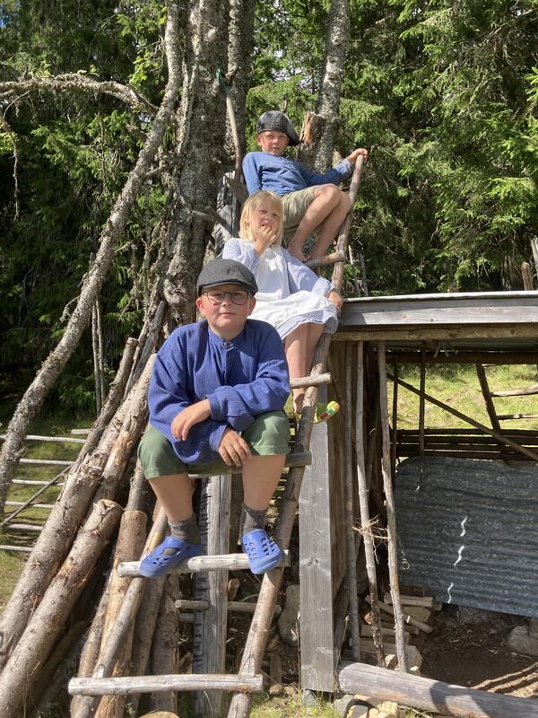 Tre barn sitter på en trestige lent mot et tre og et uthus.