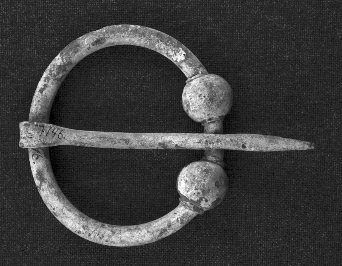 Ringspænde av bronse fra vikingetiden av typen Rygh 680 med glatte kuler paa endene av ringen, toppen av naalen dog uten kule, bare bøiet om til en hempe. Ellers er hele smykket glat og enkelt. Ringen er 6,4 cm. i største ytre tvermaal, naalen 8,6 cm. lang. Fundet under veiarbeide i en bakke paa Skeie, Kvinnherad sogn og pgd., Hordaland. Gave fra gaardbruker Johannes Skeie, Rosendal
