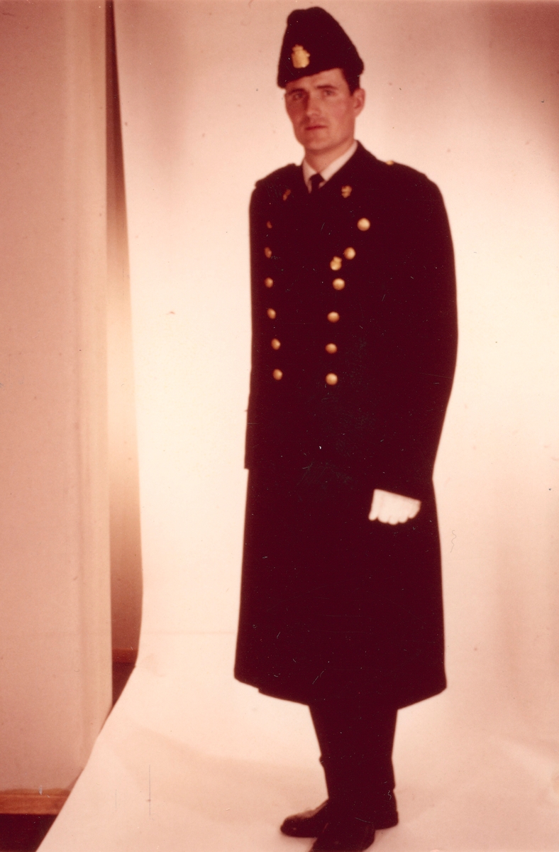 Politimann iført politiuniform, vinterlue M/1935 og helårskappe. Kappen fra 1953-reglementet og 1963-reglementet har samme snitt.