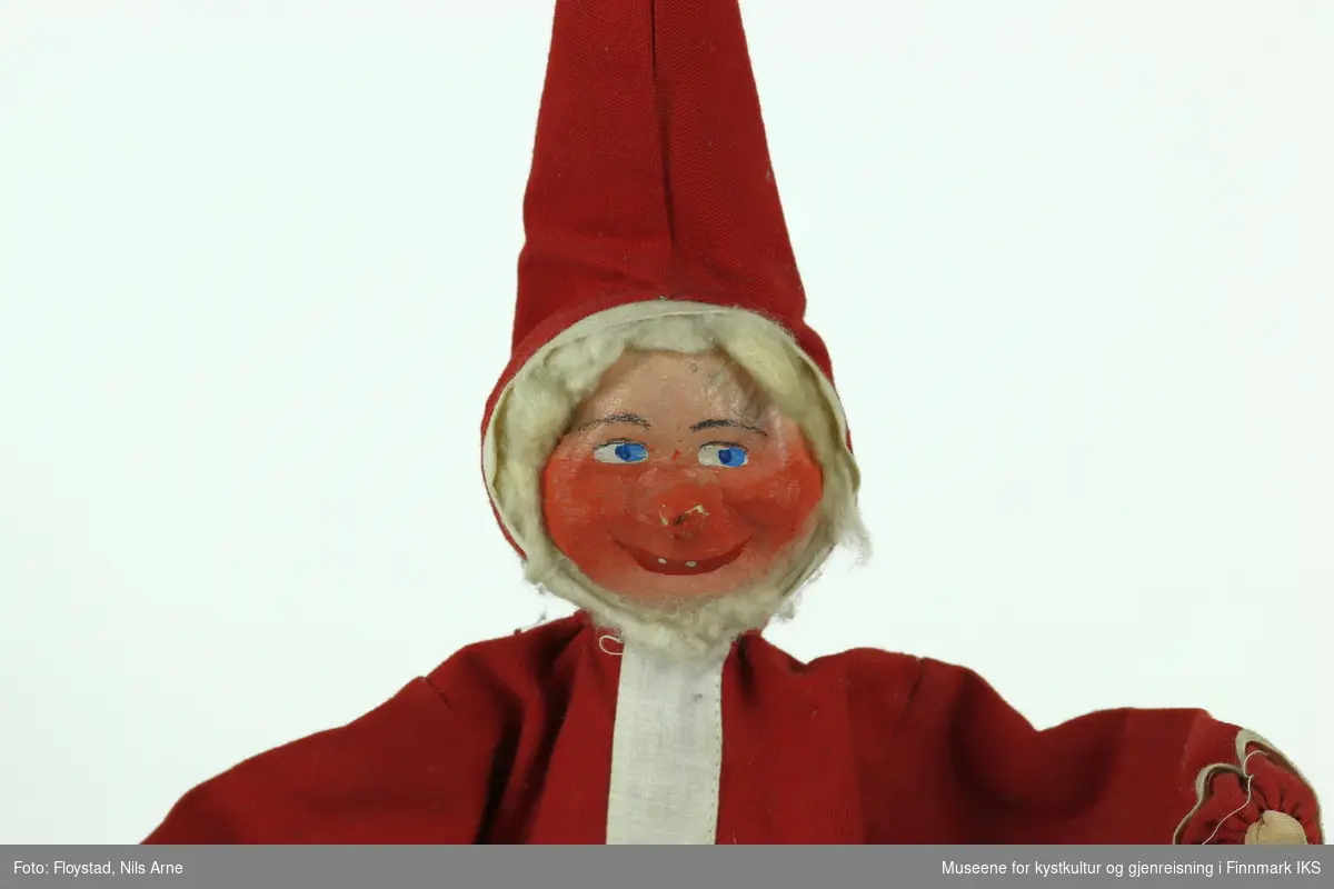 En julenisse laget av papp og tøystoff. Tøystoffet er i fargene rød og hvit, bortsett fra hendene til nissen der har tøystoffet en hudlignende farge. Ansiktet til nissen er laget av pappmasje også er det påmalt øyenbryn, blå øyner og munn. Rundt hodet er det pålimt bomull. 