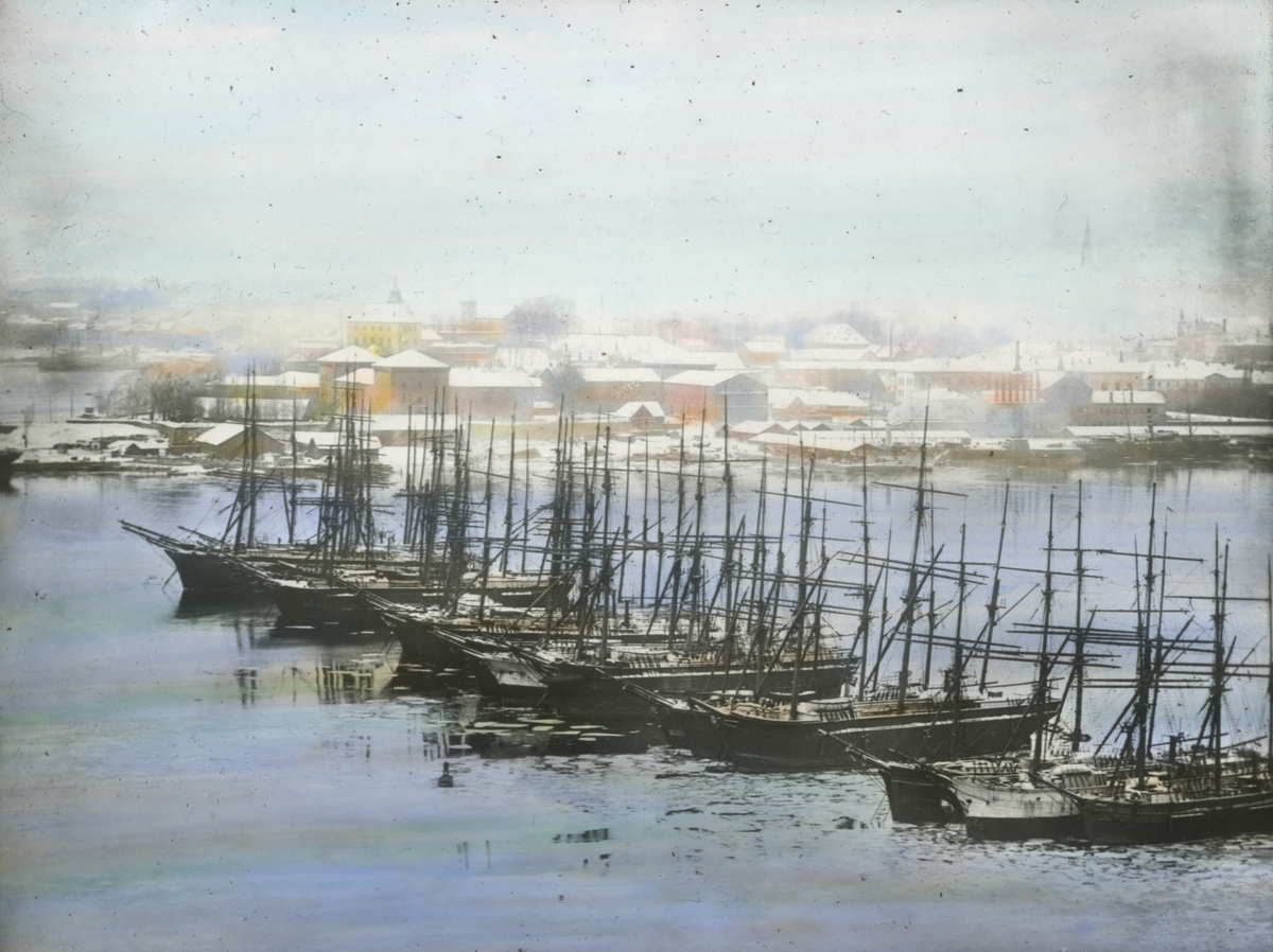Håndkolorert dias. Seilskuter ligger fortøyd i havnebassenget i Oslo havn vinteren 1901. I bakgrunnen sees Akershus festning og sentrum.