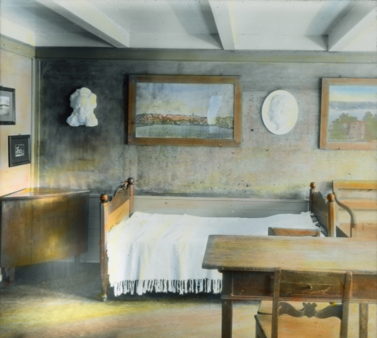 Håndkolorert dias. Interiør fra Ibsenmuseet i Grimstad.En seng står inntil vegge, i hjørnet et hjørneskap og midt i rommet står et bord med to stoler. På veggen henger en byste av Henrik Ibsen og fire malerier.