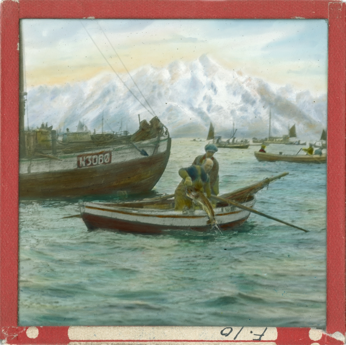 Håndkolorert lysbilde. To menn i en liten robåt drar opp en fisk fra havet. Det er flere fiskebåter på sjøen. I bakgrunnen ses fjell med snø.