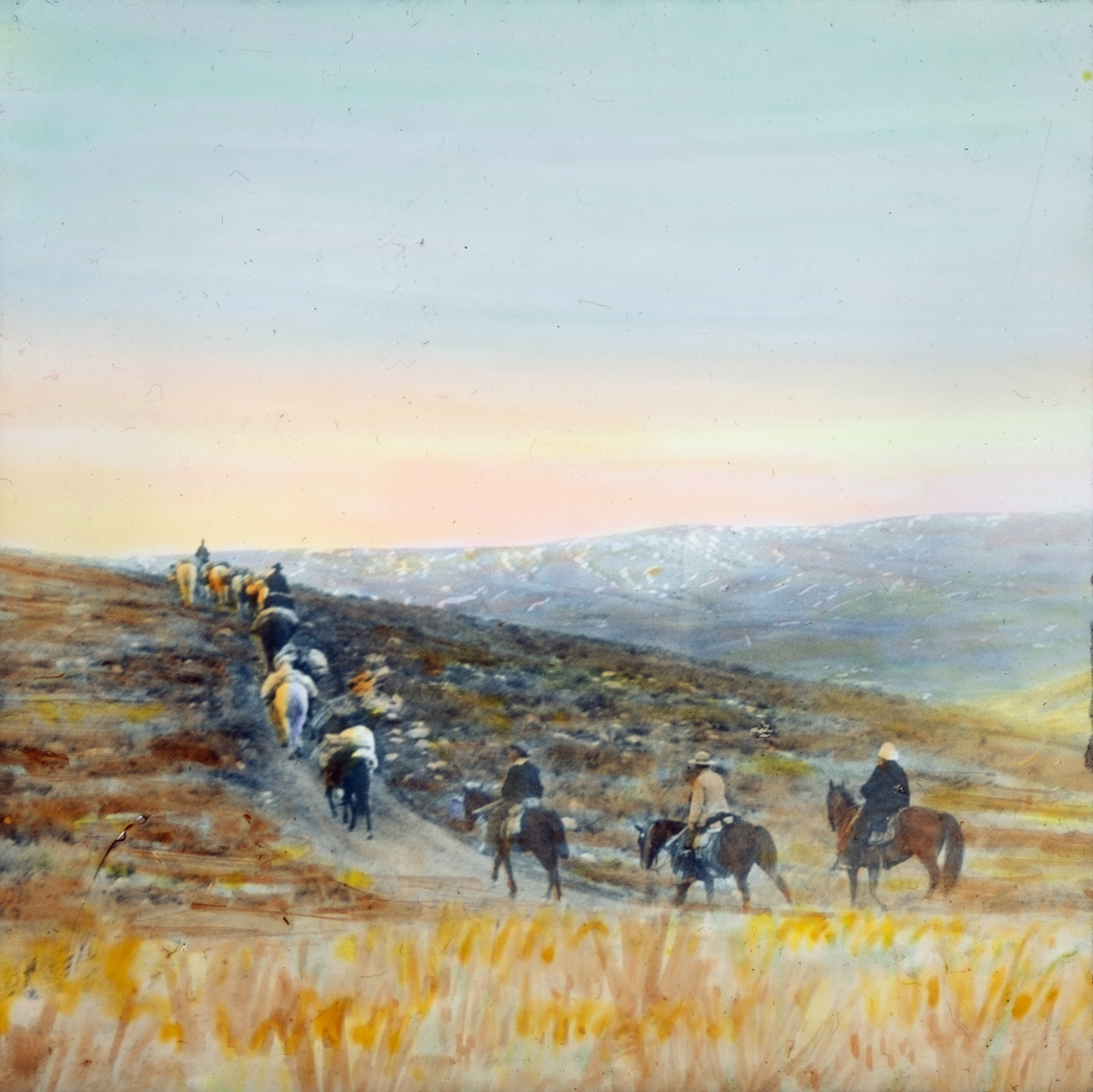 Håndkolorert dias. En gruppe menn på hest med tre pakkhester rir på en sti i et nakent slettelandskap.