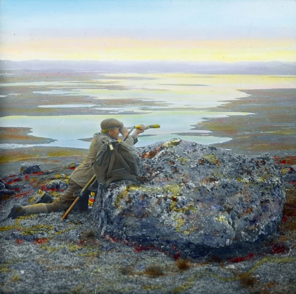 Håndkolorert dias. En mann lener seg mot en stor stein og ser utover et landskap ved Bjornesfjorden gjennom en kikkert. Ved siden av ham på steinen står en ryggsekk.
