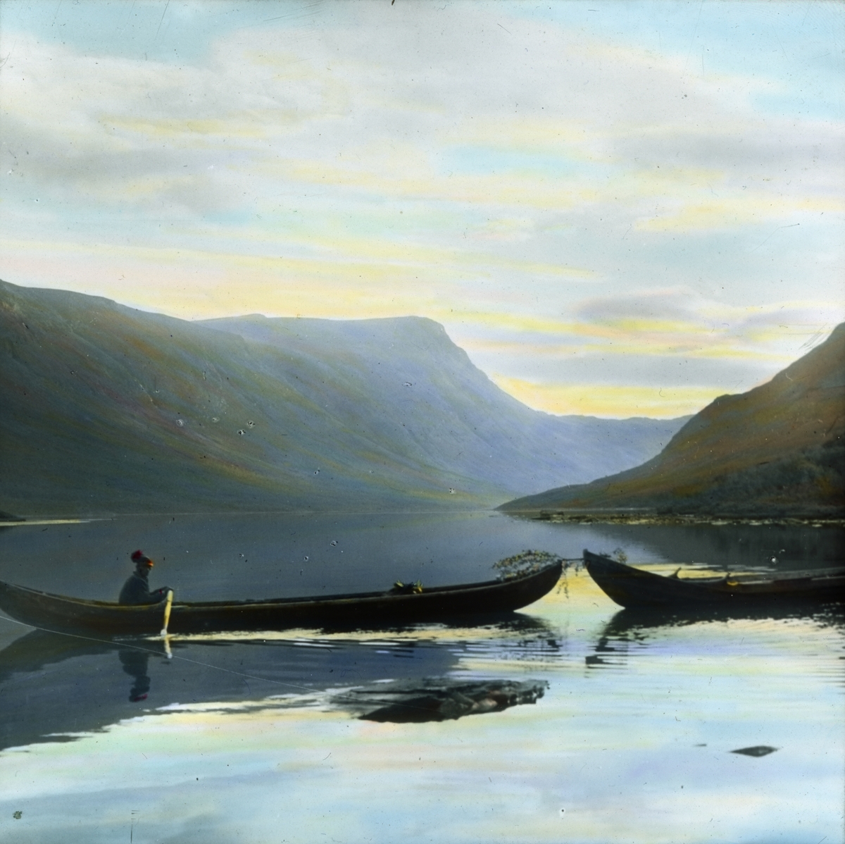 Håndkolorert dias. En mann ror og tauer en annen båt på en innsjø i nasjonalparken Pieljekaise i Sverige.