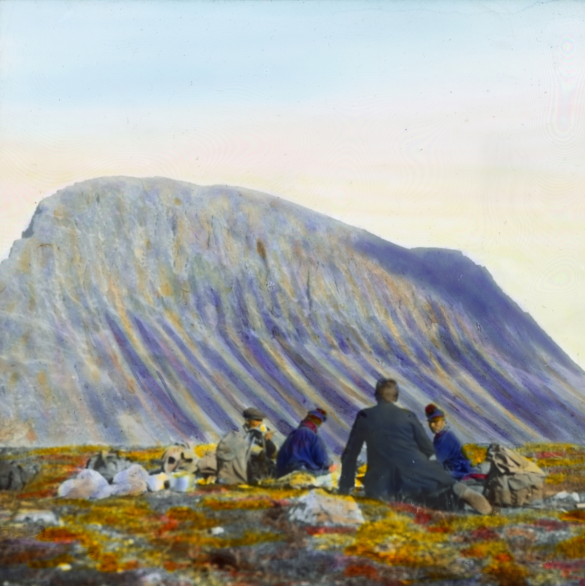 Håndkolorert dias. Fire menn, hvorav to er kledd i kofte, spiser mat ved foten av fjellet Laidopakto.