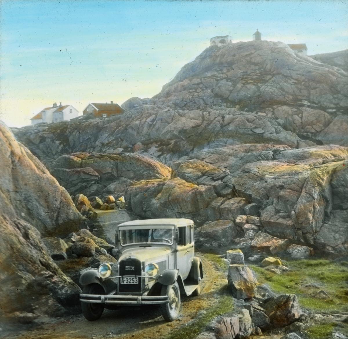 Håndkolorert dias. En bil, Citröen 1929-SIX, kjører mellom knauser i landskapet ved Lindesnes fyr. I bakgrunnen skimtes fyrvåkterboligen.