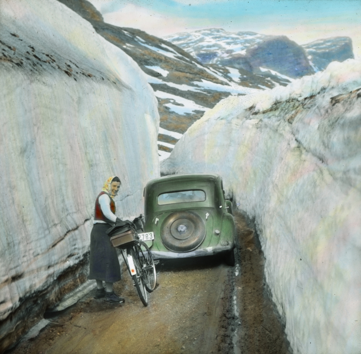 Håndkolorert dias. En kvinne med sykkel står bak en bil blant høye snefonner i et høyfjellslandskap.