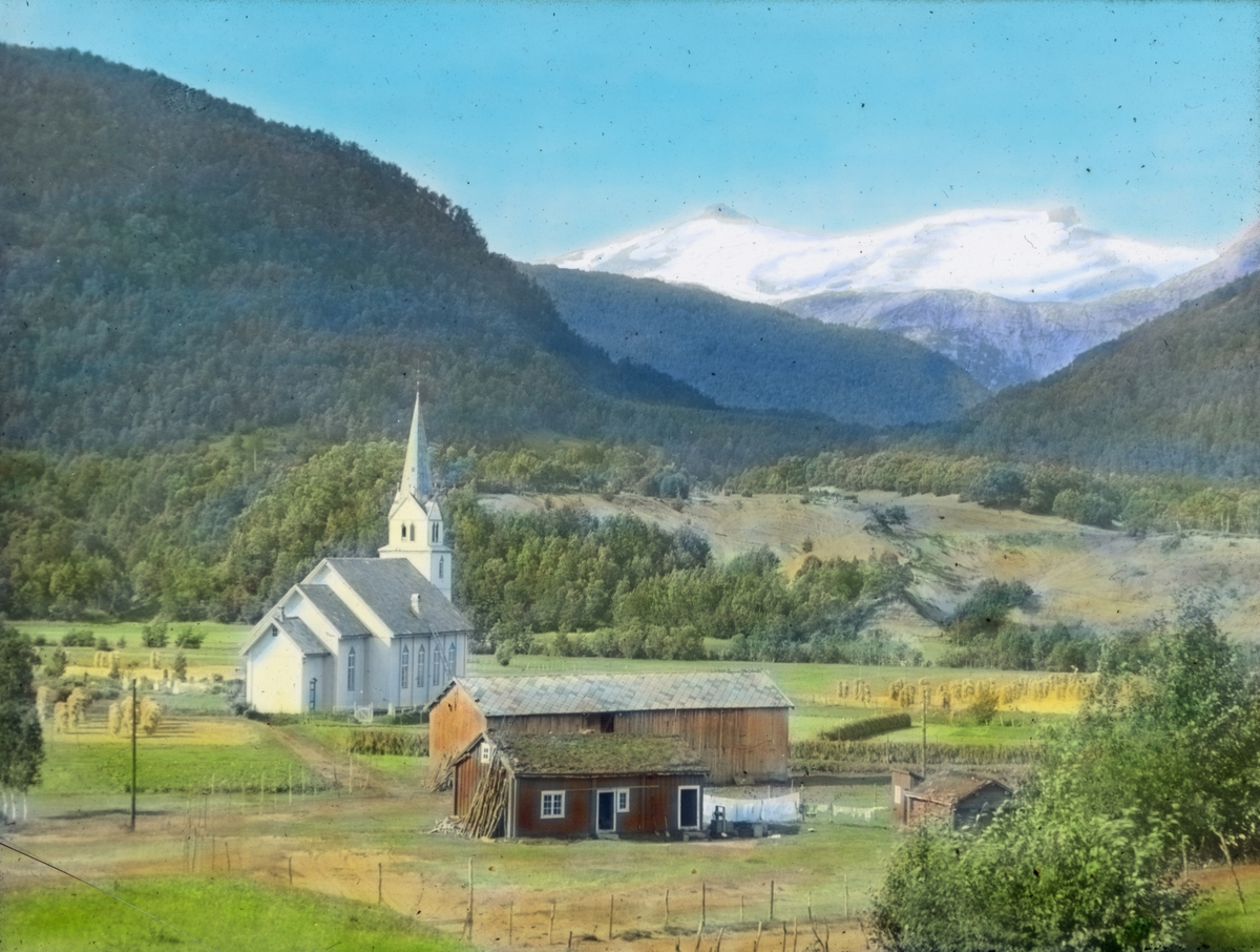 Håndkolorert dias. En hvitmalt kirke i et jordbrukslandskap. I bakgrunnen sees bratte åser og snødekte fjell.