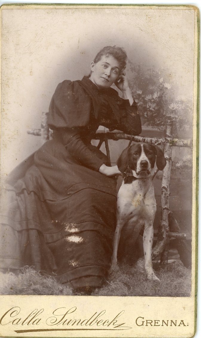 Kabinettsfotografi av en okänd kvinna som sitter vid ett enkelt staket av trä i en ateljé. Hon har en hund vid sin sida.