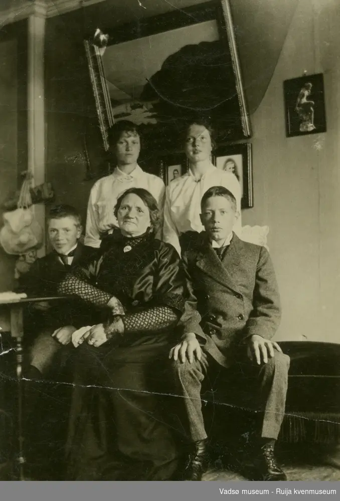 Gruppebilde av Olsen familien i stuen. Den ene gutten har dobbeltspennt dress, damen i midten har Kamelia -smykke rundt halsen. Fra venstre bak: Lilly Olsen og Borghild Olsen. I midten foran: Anna Olsen (født Helgesen). Gutten foran til høyre er antakelig Erling Olsen. Ca 1910.
