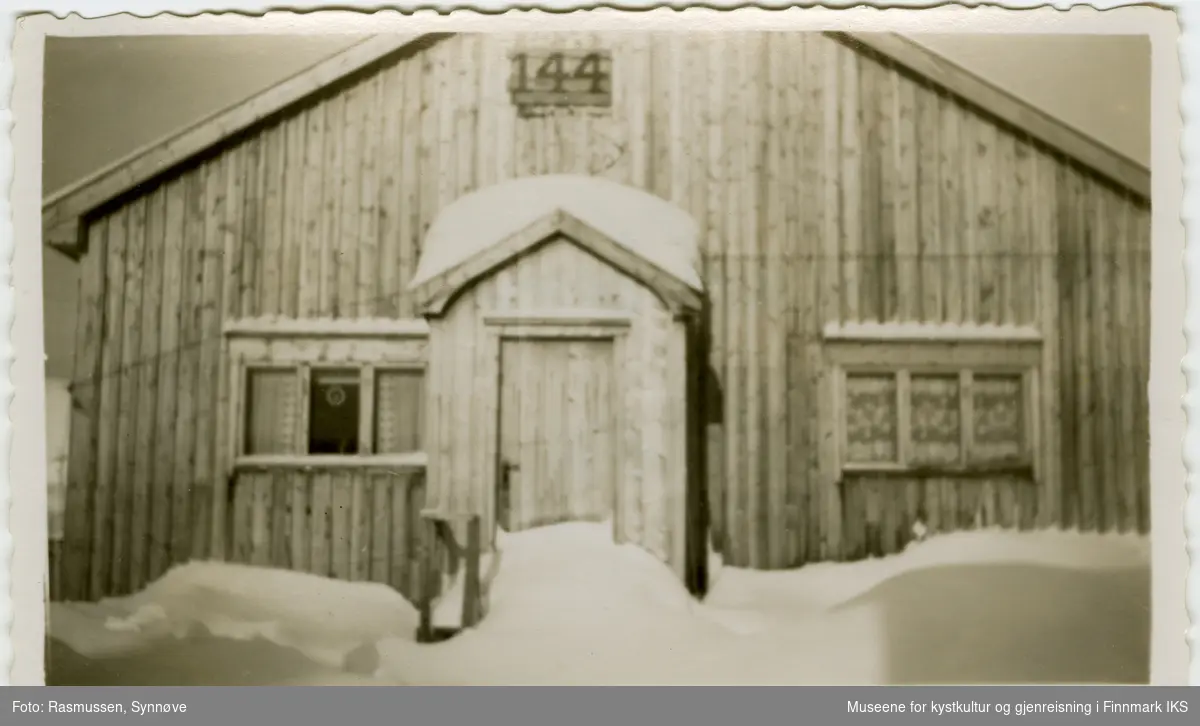 Bildet viser brakke 144 i Hammerfest i gjenreisningstiden. Snøen ligger høyt. Brakka har to tredelte vinduer, et på hver side av inngangen. Brakkenummeret er montert med store sifre på en ventil som fører til loftet som ble brukt til lagring.