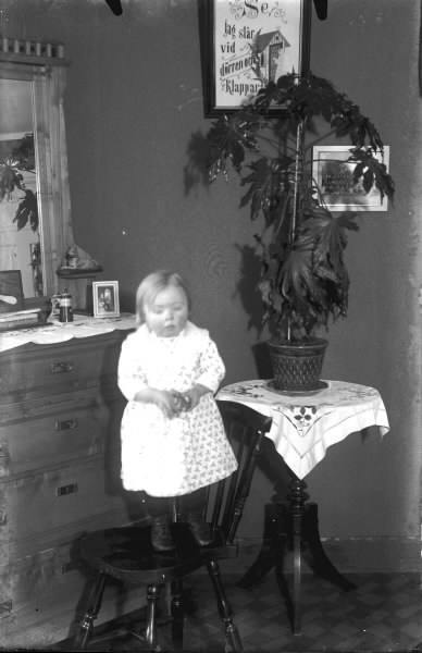 Litet barn står på en stol vid en spegelbyrå och ett pelarbord med en hög växt. På väggen hänger en tavla med en ängel vid ett hus och texten: Se jag står vid dörren och klappar.