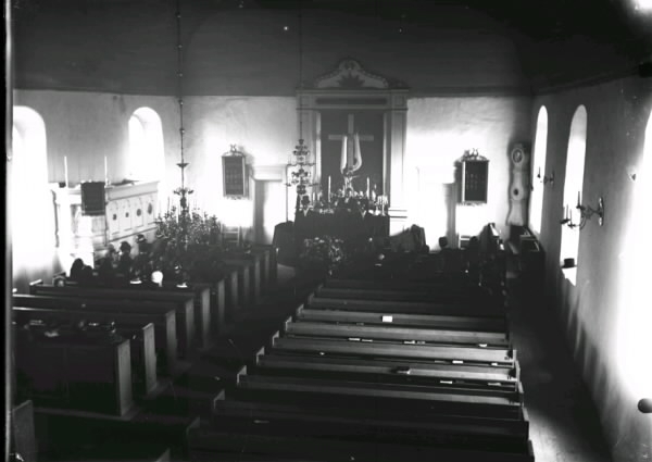 Interiörbild från Drängsereds kyrka vid en begravning. Fotot är taget från orgelläktaren mot koret. Vid kistan står män i höga hattar. Till vänster ses ett skrank som döljer trappan upp till predikstolen.