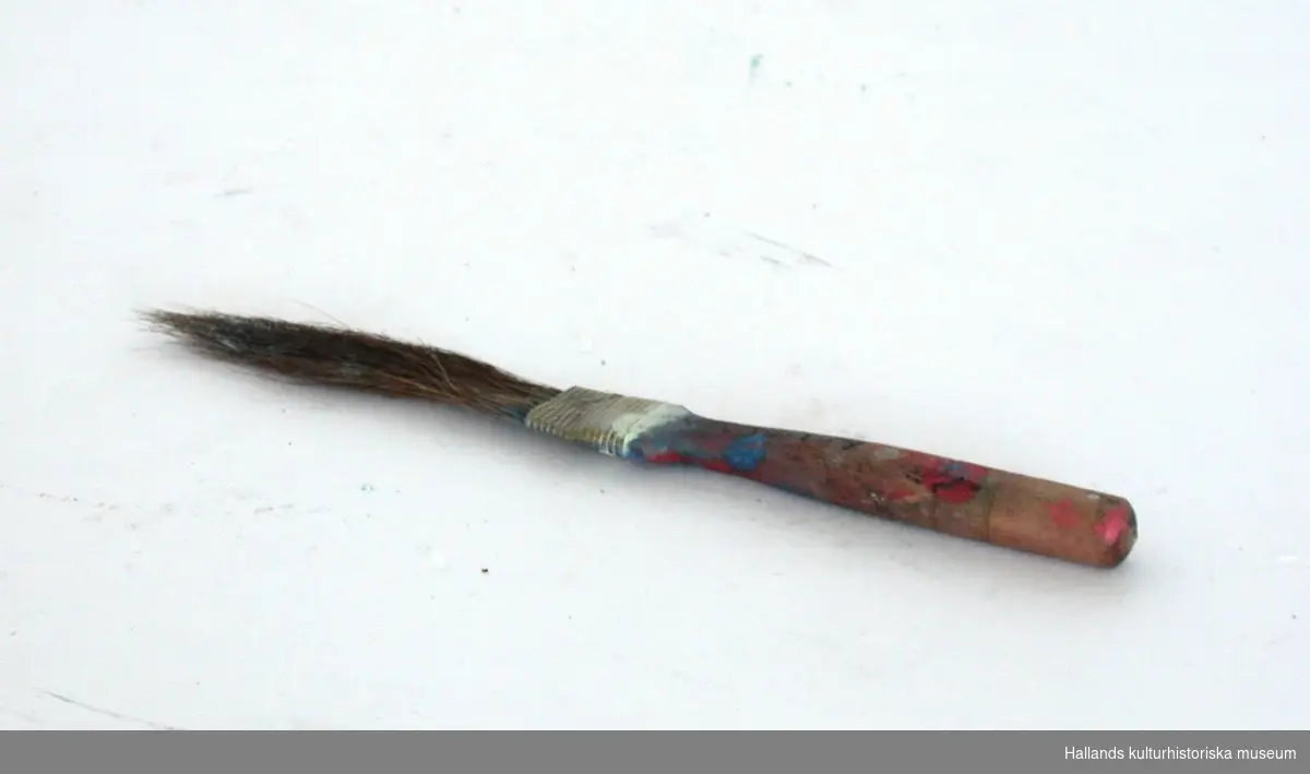 Liten pensel med träskaft. Skaftet smalnar av där "penselhåren", är fastklämda med en metallbit. Blå, röd och vit målarfärg finns på skaftet.