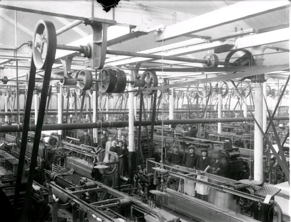 Fabriksinteriör där maskinerna står tätt och drivs med remdrift. Några arbetare står bland maskinerna.