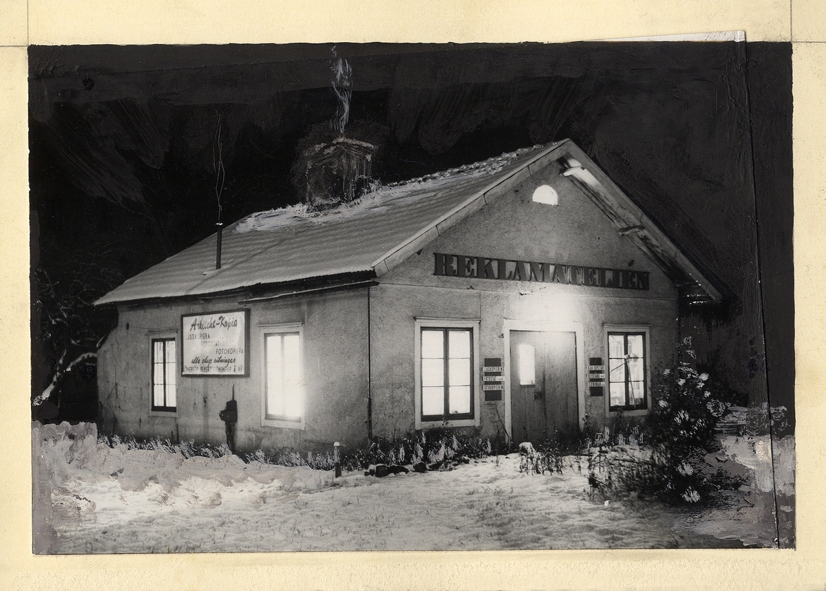 En stuga i vinterskrud med texten: "Reklamateljén" på gaveln.
På långsidan syns en annan skylt med text: "Arkitekt-Kopia, Ljuskamera..."