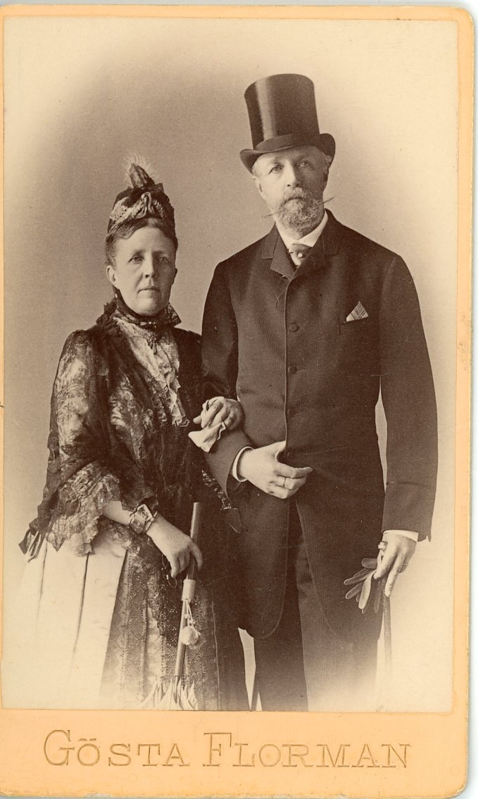 Kabinettsfotografi av en högtidsklädd kvinna och man, de håller armkrok. Kvinnan håller ett parasoll och mannen bär hög hatt.