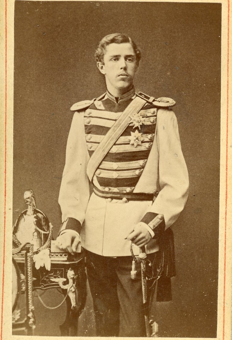 Kabinettsfotografi av en ung man i militär uniform, med vänster hand på värjan och en kask på ett bord intill.