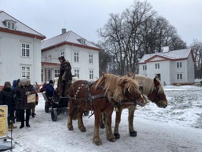 Et tospann med hester tar på passasjerer foran Eidsvollsbygningen. Lett snødryss på bakken og på taket til Eidsvollsbygningen og søndre sidebygning i bakgrunnen.