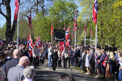 Et 17.mai-tog ankommer Eidsvollsbygningen. Vi ser en politi i uniform og speidere med flagg i flaggborg først i toget. Langs kanten står masse mennesker og ser på. Mange kledd i bunad, mange norske flagg i bildet.