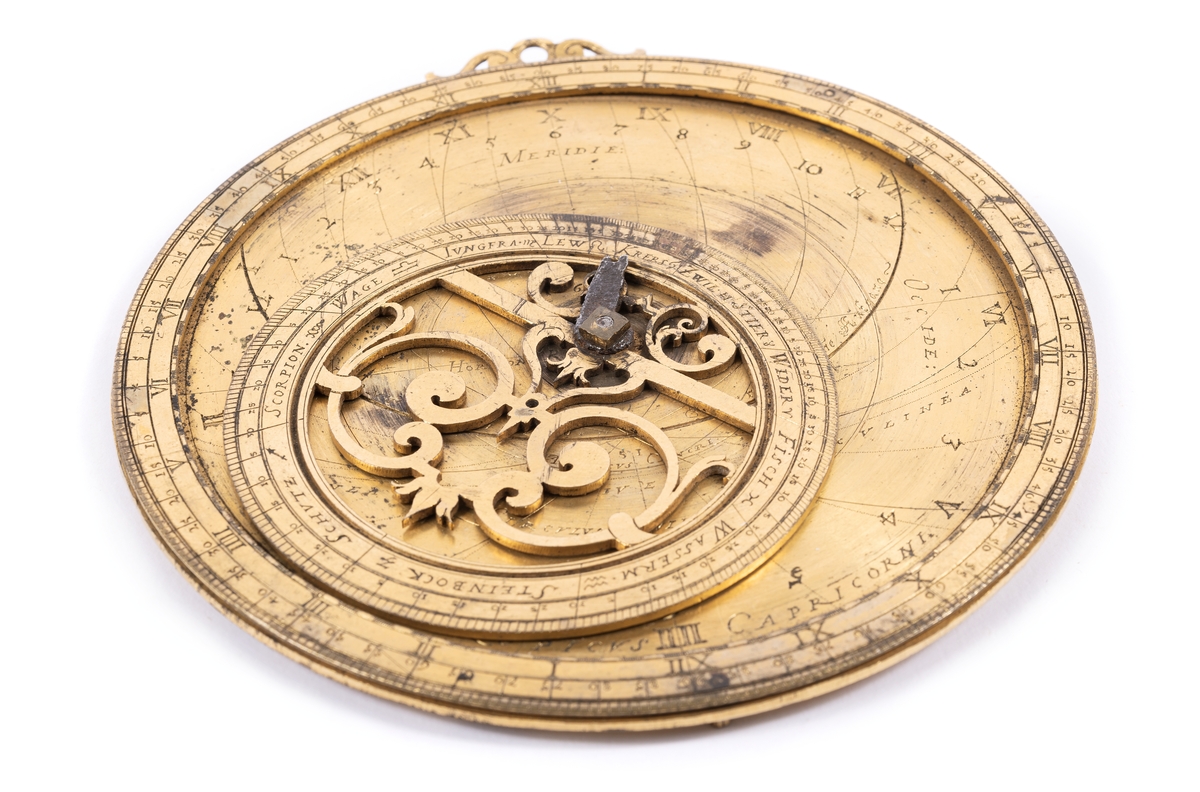 Astrolabium av förgylld metall, med inskriptioner på tyska. På framsidan inom cirkelrund kant, excentrisk rörlig genombruten zodiakring, samt fragment av diopterlinjal av järn. På baksidan månkalendarium med två rörliga skivor.