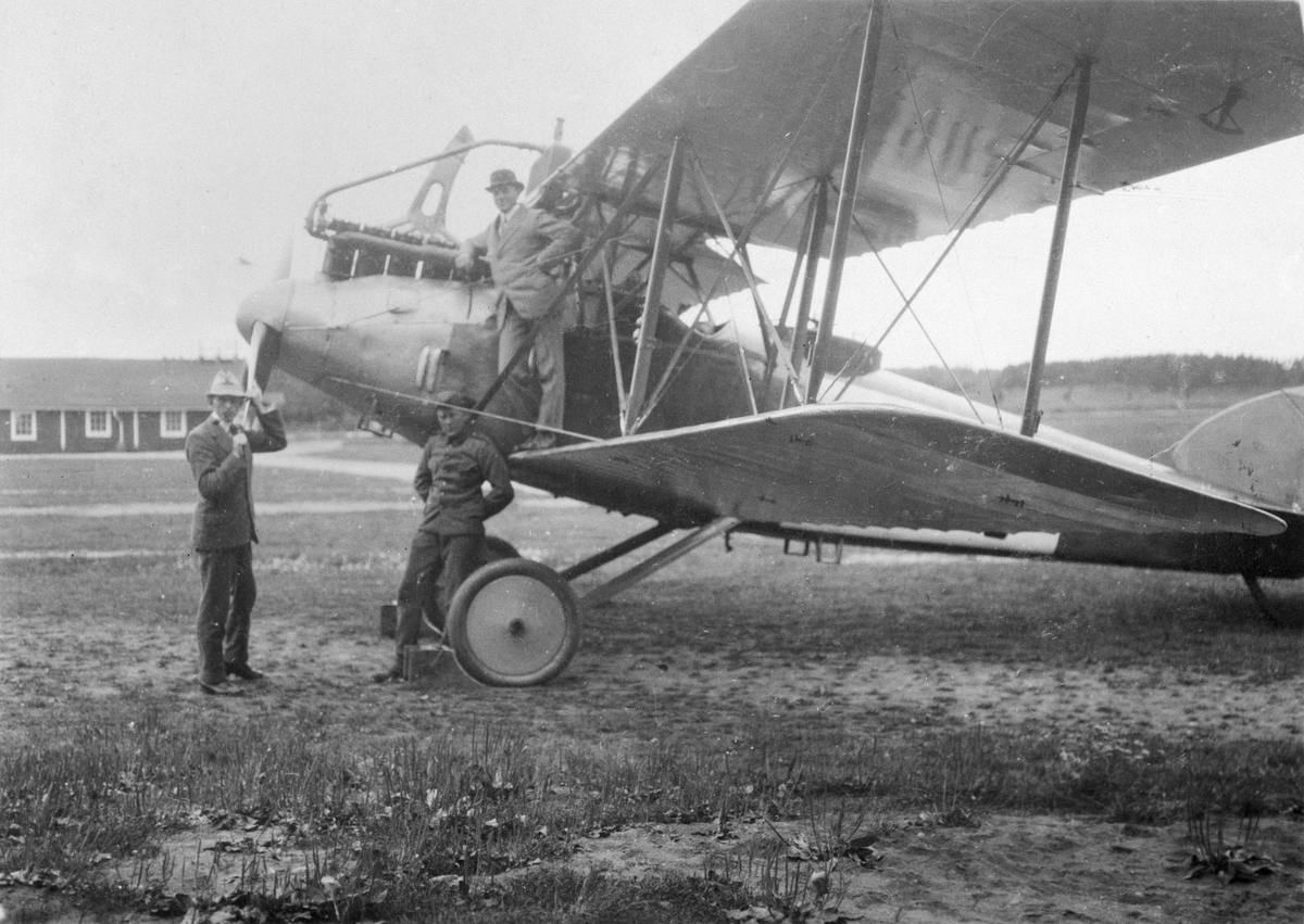 Två civilklädda och en militär poserar vid flygplan FVM S 18 på Flygkompaniets tygverkstäder på Malmen, omkring 1920. I bakgrunden syns filarverkstaden.