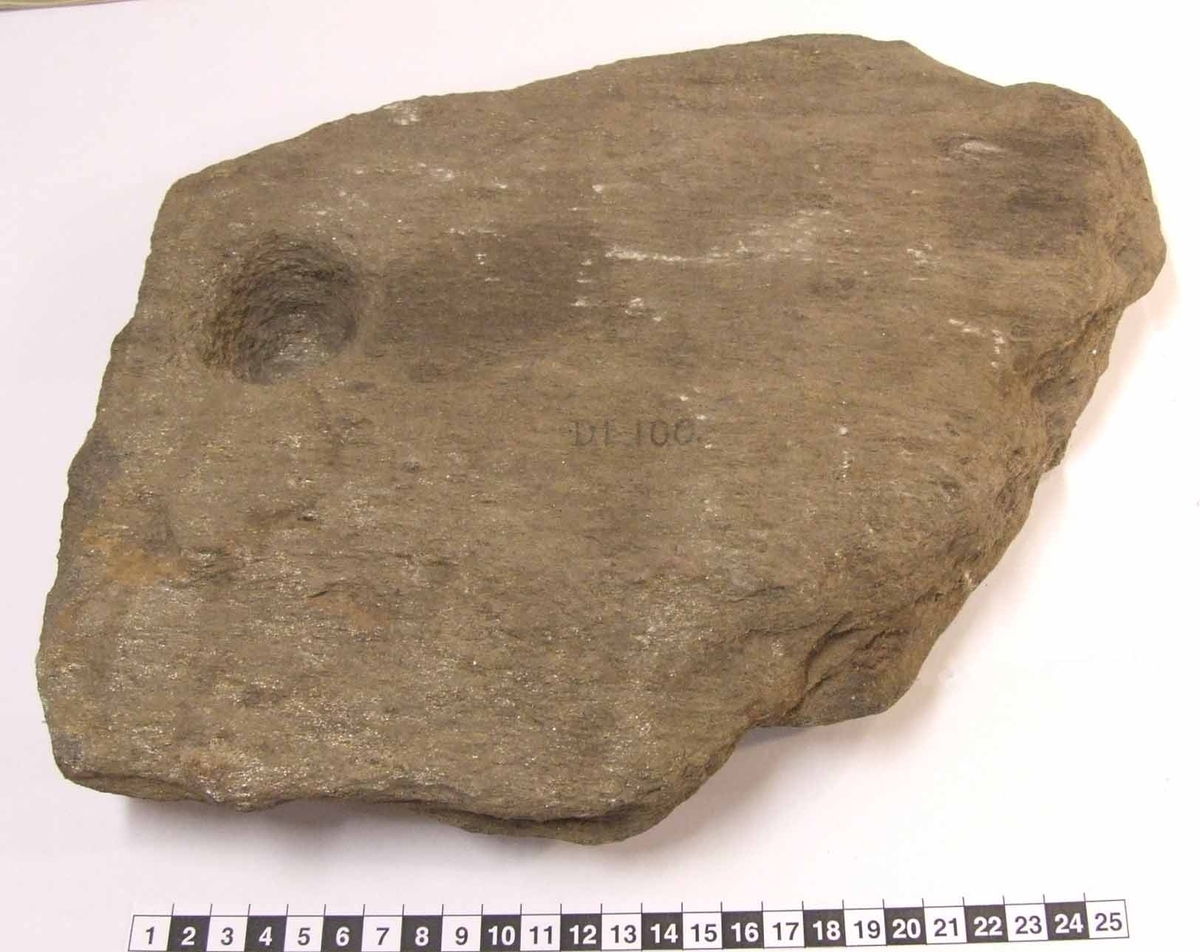 Älvkvarnsten funnen i gravhög. Flat sten av glimmerskiffer.
I ena kanten är en älvkvarn/skålgrop som är 51 mm i diameter och 25 mm djup, med grov yta.