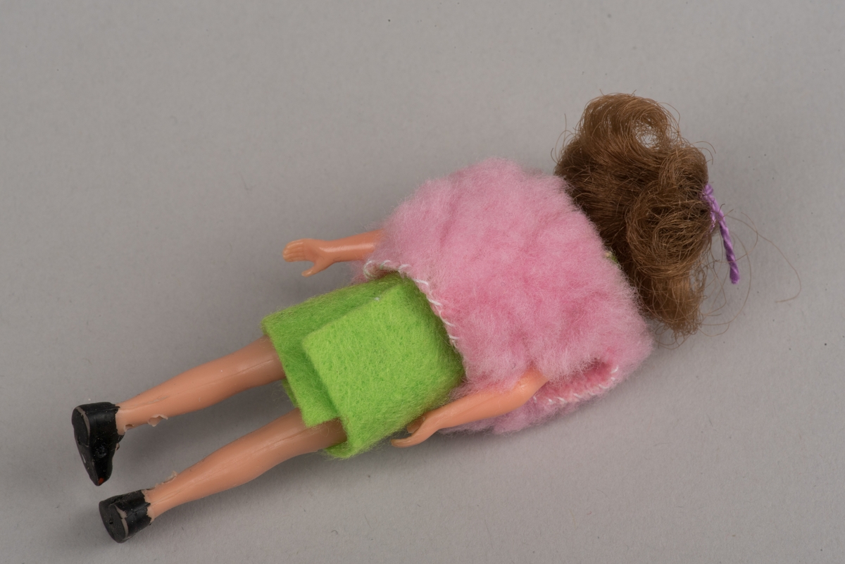 Flickdocka för dockskåp, troligen tillverkad av Lundby leksaksfabrik, i plast.
Dockan är klädd i pålimmad ärmlös klänning av grön filt och en rosa ärmlös väst. På fötterna ditmålade svarta skor.
Håret är ihophållet av en lila tråd i tofs.