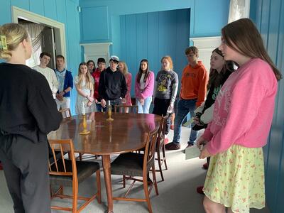 En gruppe ungdomsskoleelever i sommerklær er på omvisning i Eidsvollsbygningen, her står di i konstitusjonskomiteens værelse.