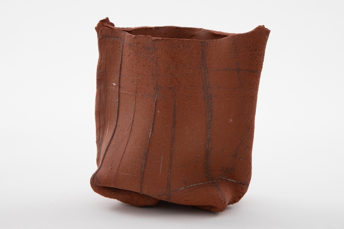 Matt rødbrun vase i steingods med grunnform som en spiss oval og rette vegger som er klemt litt inn nederst. Leireplatene overlapper hverandre på sidene og danner en hanklignende kant. Tekstur i overflaten. Åpen munning.