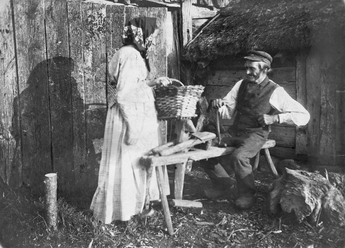 Henrik i Björsgård (granne med Severin) och en kvinna med korg som han just tillverkat (se bildnr E5819_1). Han sitter på en "träskomärr" i vinkeln mellan två huskroppar med skiftesverksstommar.