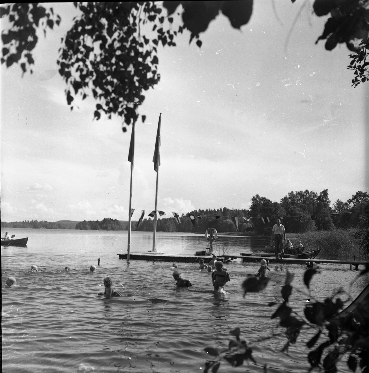 Några barn simmar i en sjö. Det står vuxna på en brygga och tittar på. Två flaggstänger står längst ut på bryggan. En roddbåt skymtar i utkanten av bilden.