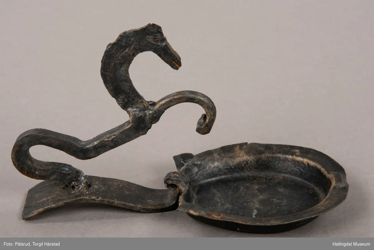 Et askebeger med håndtak i form av en steilende, stilisert hest. Hesteskoformet askebeger. Laget i smijern, og er dekorert med sort og påpusset gull. 
