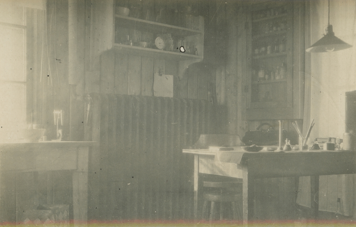 Fotografi från expedition till Spetsbergen. Interiörbild från trähus med motiv av rum med två bord, varav ett skrivbord, element hyllor med diverse små flaskor, taklampa och fönster.