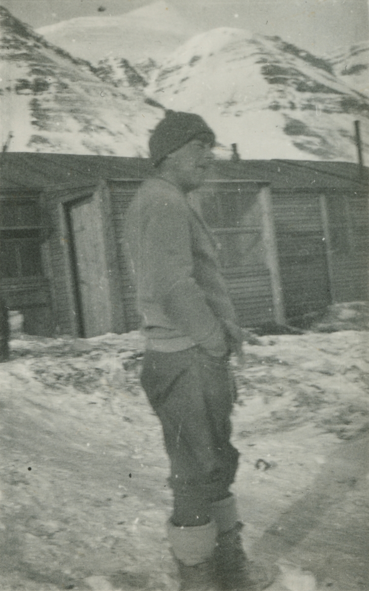 Fotografi från expedition till Spetsbergen. Motiv av man som står i snön framför ett trähus. I bakgrunden syns ett snötäckt berg. Forografiet troligtvis taget vid Sveagruvan.