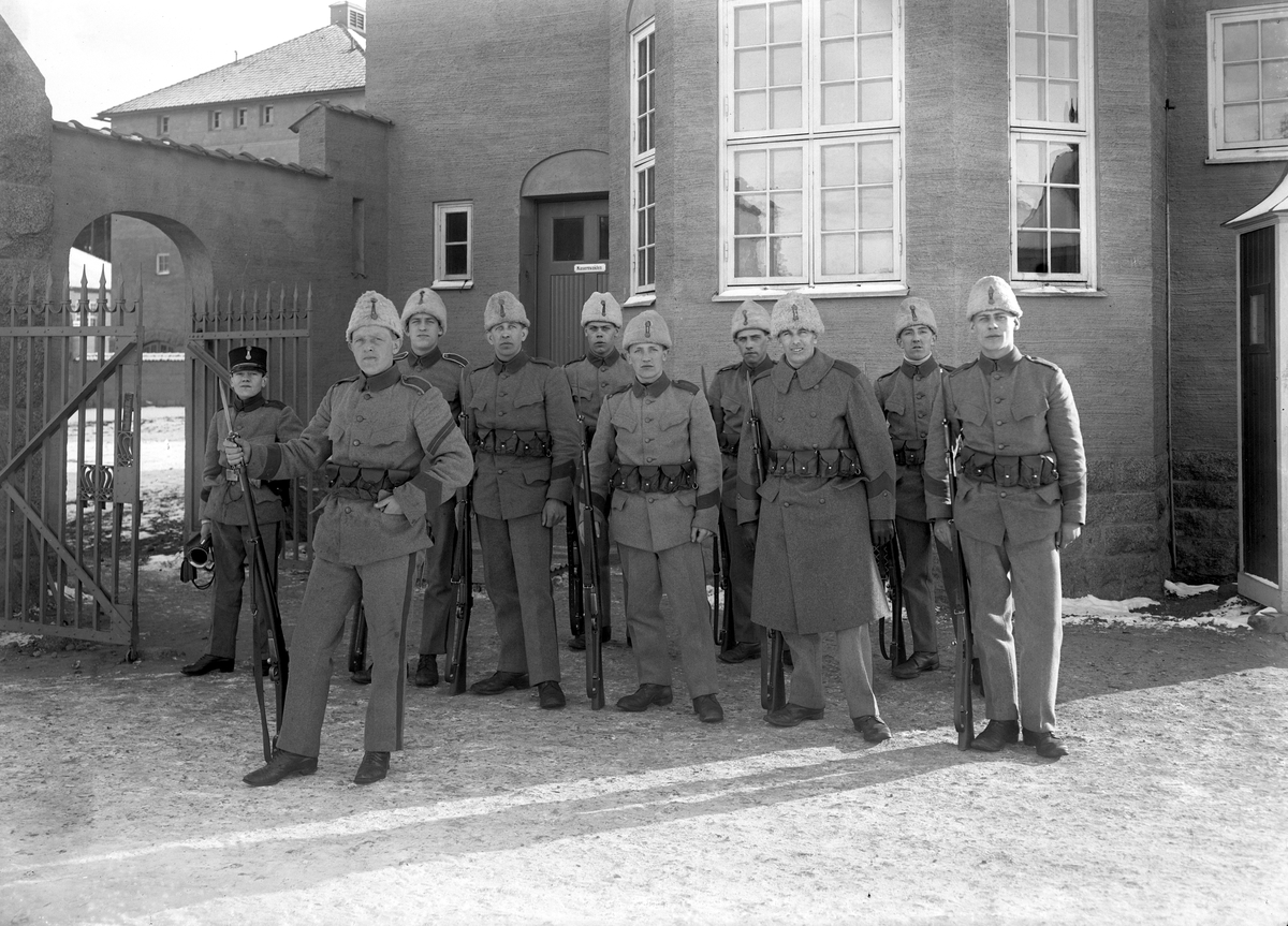 En grupp infanterister, möjligtvis i vakttjänst, samlade för fotografering invid den gemensamma vakt- och arrestlokalen för Linköpings bägge infanteriregementen. De är iförda uniformer av modell 1910 men den odaterade bildens tillkomst knyts till påföljande decennium.