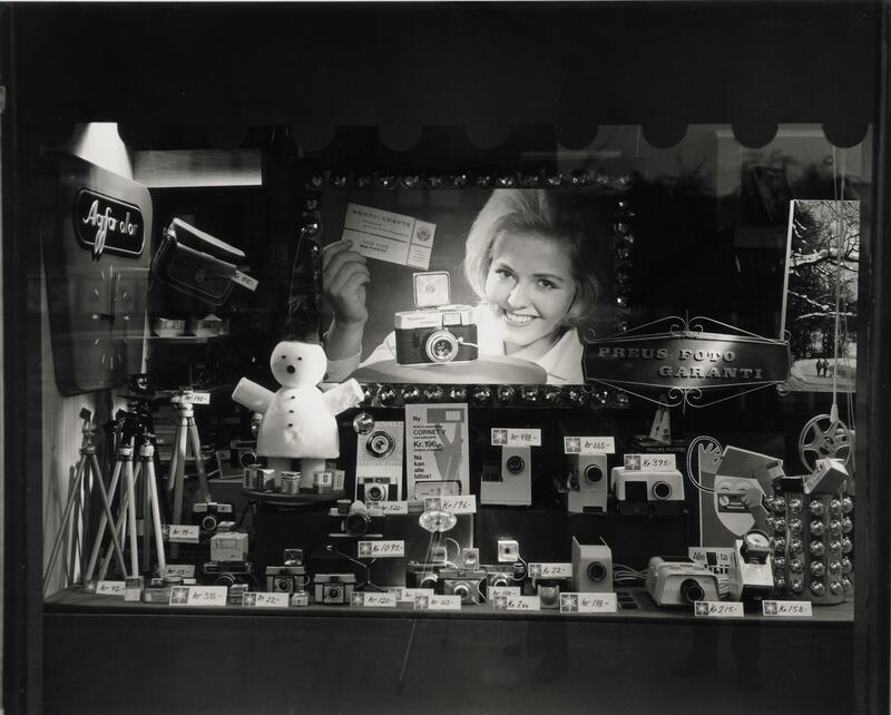 Butikkvindu med kameraer til salgs og et fotografi av en smilende kvinne. Svart-hvitt fotografi.