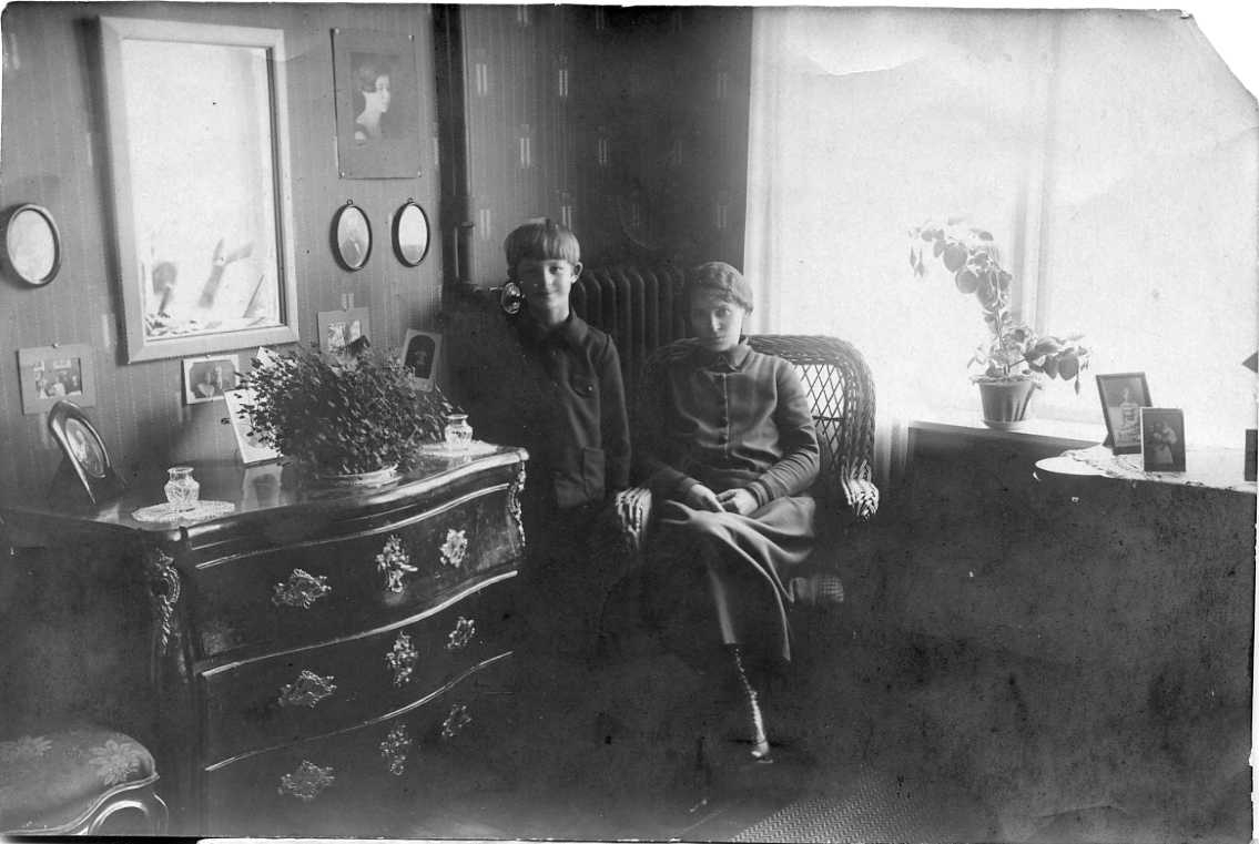 En pojke står intill sin mamma som sitter en rottingstol framför ett fönster. På väggen till vänster om pojken hänger ett porträtt av hans storasyster.