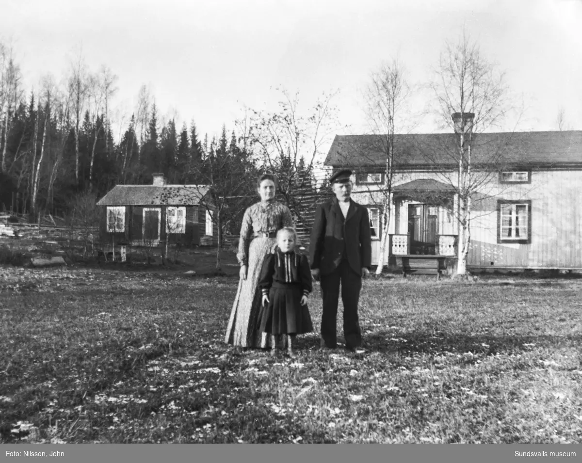 En kvinna, en man och en flicka framför en ljusmålad mangårdsbyggnad med brukstuga intill. Bakom husen skymtar en storhässja.