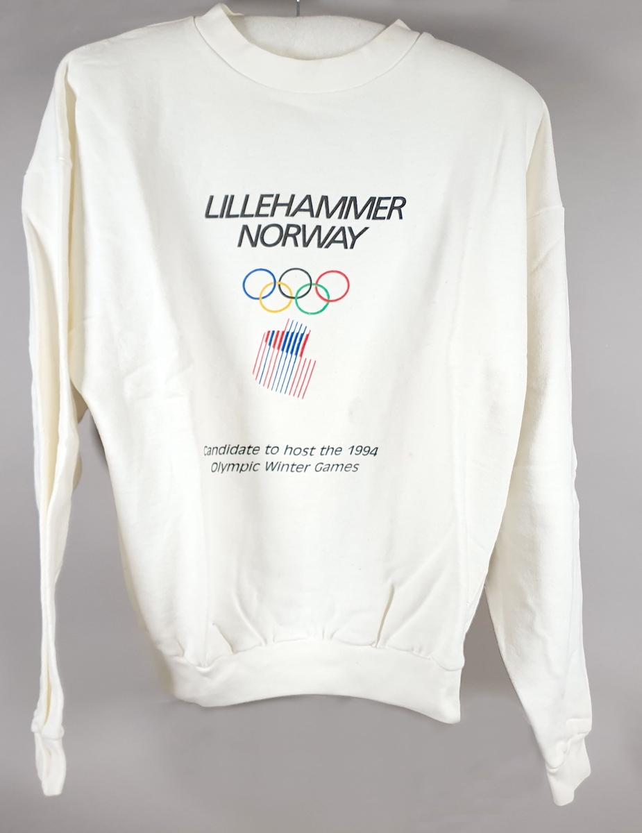 Hvit genser med lange ermer med logo for søkerbyen Lillehammer-OL 1994, og olympiske ringer i farger.