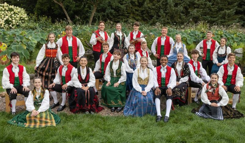 23 unge amerikanske folkedansere, både gutter og jenter kledd i norske bunader poserer på et grupppebilde, sittende på en grønn plen med grønne busker bak seg.