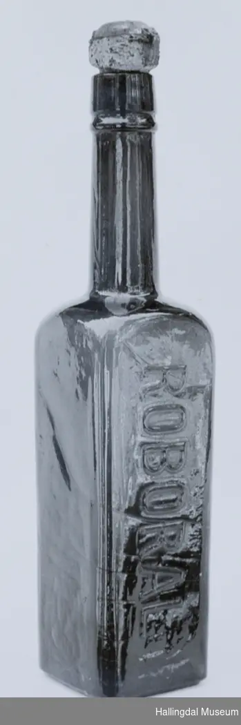 Selve flasken rektangulær med kvadratisk tverrsnitt. Tuten er sylindrisk.
6/6-68. Arne L. Christensen.
