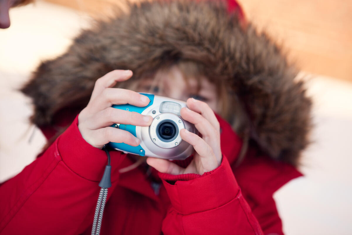 Et barn med vinterjakke holder et kamera opp til øynene og peker det mot oss som ser på bildet.