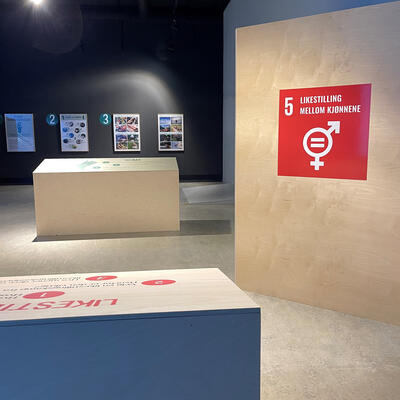 Foto fra utstillinga "Menneskeskapt" viser to utstillingselement på gulvet, fire plakater på veggen i bakgrunnen og en vegg med en stor logo på. Logoen er for FNs bærekraftsmål nummer 5 - likkestilling mellom kjønnene.