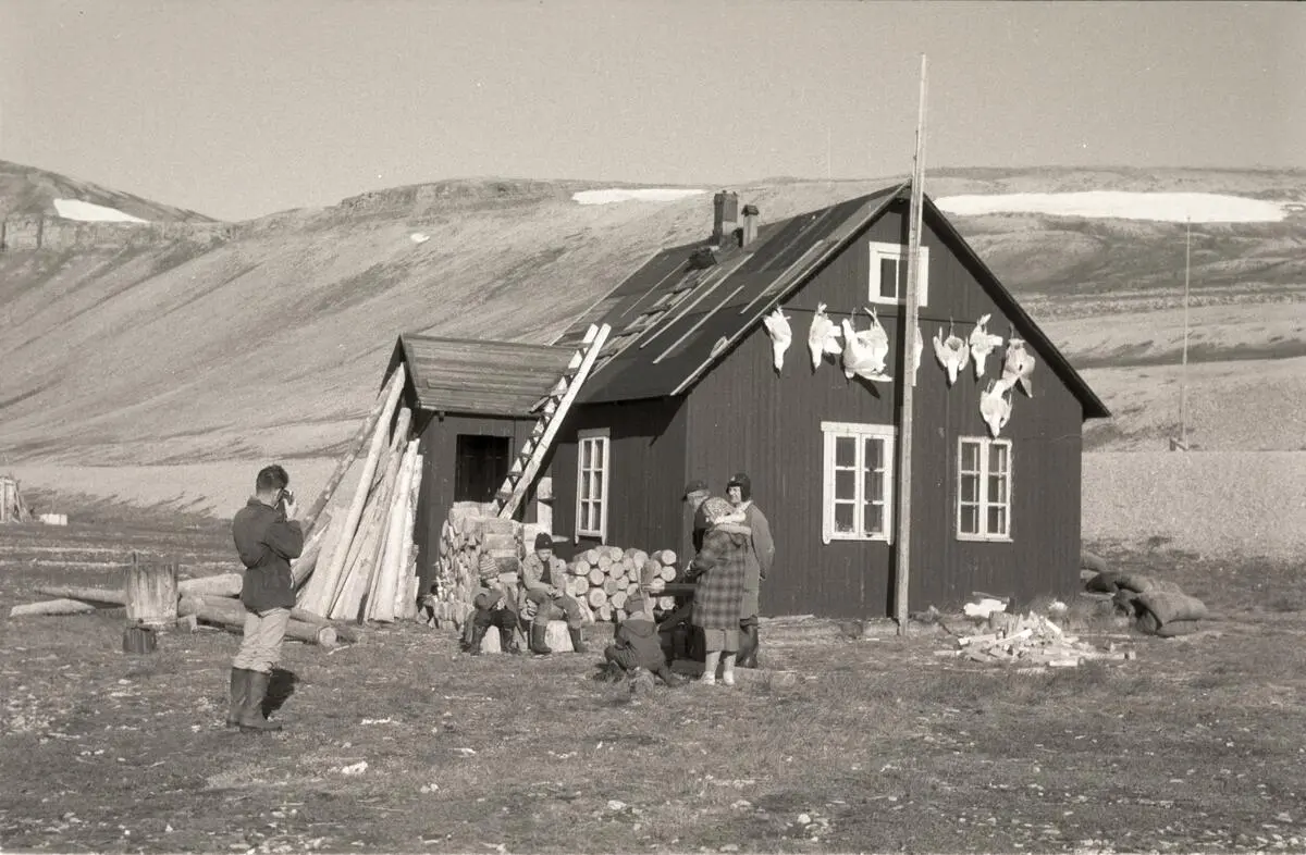 Bildet viser Villa Fredheim. Huset er omgitt av fjord, fjell og en vid dal. Foran huset er det flere mennesker som er på besøk hos Nøis i 1969. Det er store stabler av ved foran huset. Det ser velstelt og fint ut.